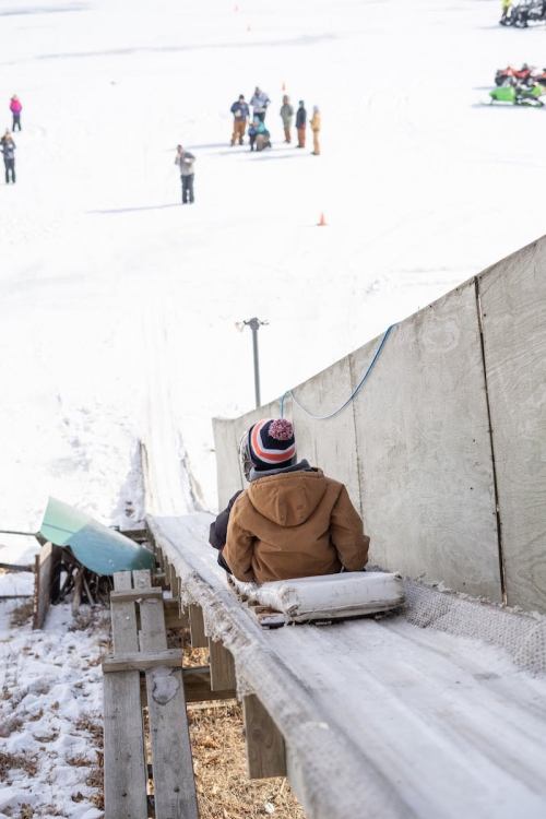 A child sits on a sled at the top of a snowy slide, ready to go down while others enjoy the winter scene.