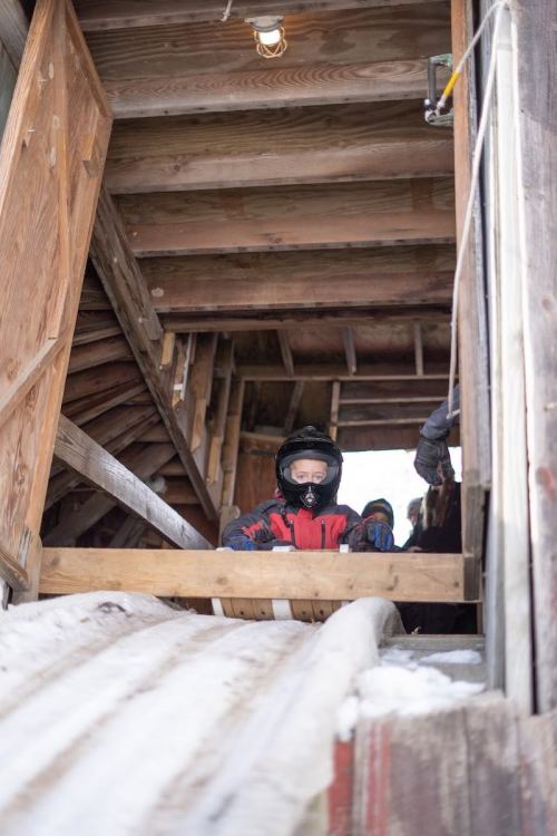 A child in winter gear sits on a sled at the top of a snowy ramp, ready to slide down. Wooden structure in the background.