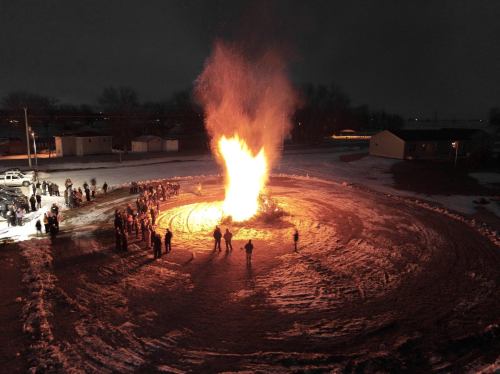 A large bonfire illuminates a snowy area, with a crowd gathered around it at night.