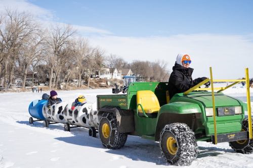 A green tractor pulls a colorful sled on a snowy landscape, with two children inside wearing winter hats.