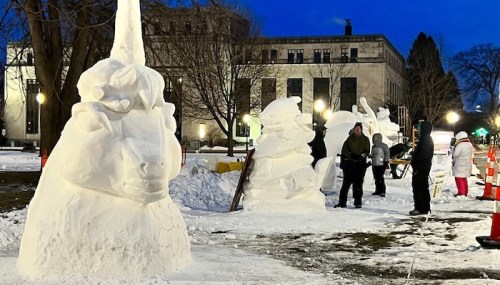 People sculpting large snow figures in a park during twilight, with snow-covered ground and trees in the background.