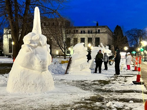 Snow sculptures, including a unicorn, are displayed in a park at dusk, with people working nearby.