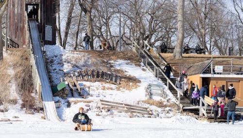 A snowy hill with a slide, people enjoying winter activities, and children playing near a wooden structure.