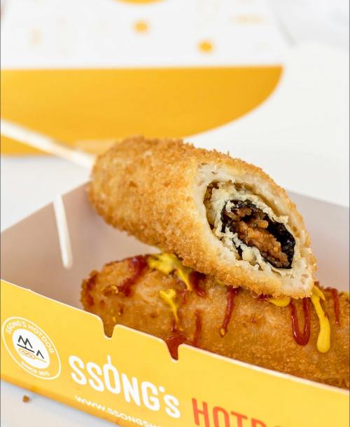A close-up of a crispy, golden hot dog on a stick, partially bitten, with ketchup and mustard drizzled on top.
