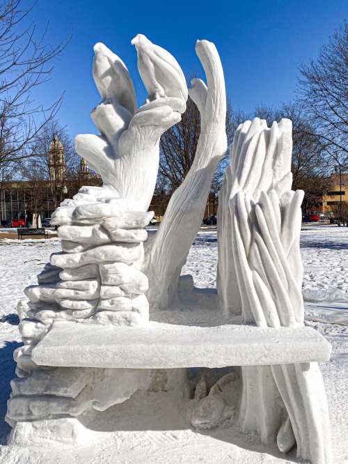 A snow sculpture resembling a bench with stylized tree branches and rocks, set against a clear blue sky.