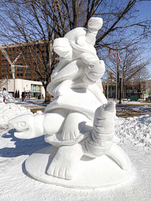 A snow sculpture of three stacked turtles in a snowy park, with trees and buildings in the background.