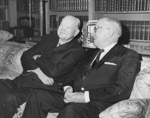 Two men in suits sitting on a couch, smiling and engaged in conversation, with bookshelves in the background.