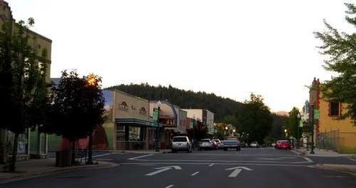 A quiet street lined with colorful storefronts and trees, leading towards a forested hillside at dusk.
