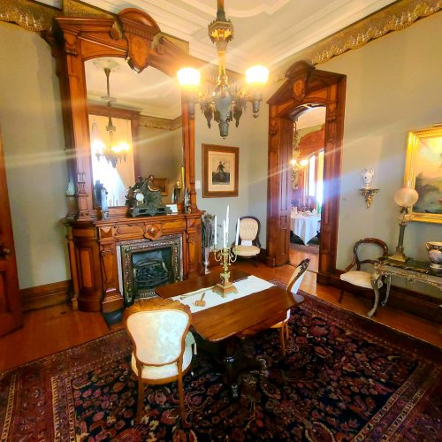 A vintage dining room with ornate wooden furniture, a chandelier, and decorative wall art, featuring a rich rug.