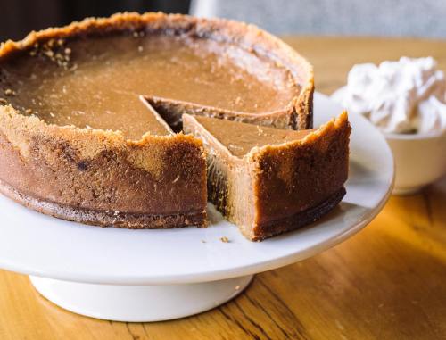 A slice of rich chocolate cake on a plate, with a whole cake in the background, set on a wooden table.