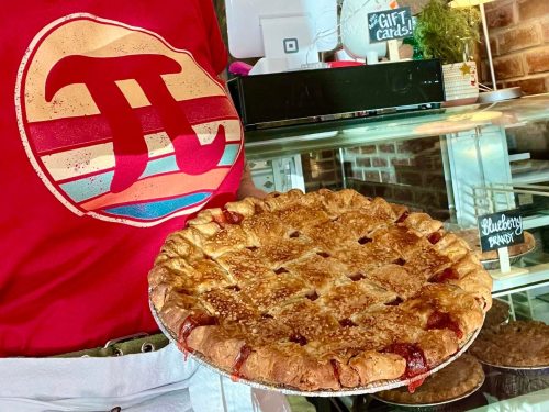A person in a red shirt with a pi symbol holds a freshly baked pie in front of a display case.