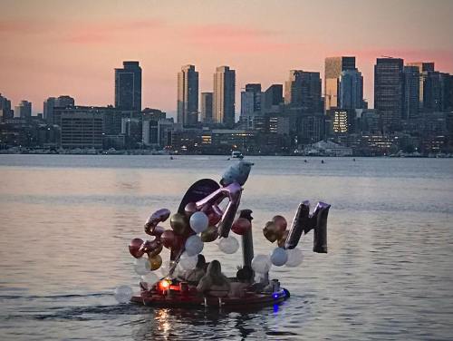 A decorated boat with balloons floats on water, with a city skyline and sunset in the background.