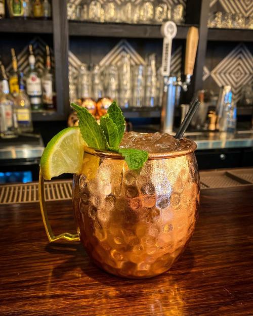 A copper mug filled with a cocktail, garnished with mint leaves and a lime wedge, sits on a wooden bar.