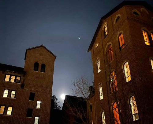 Historic brick buildings illuminated at night, with a full moon visible in the background. Snow on the ground.