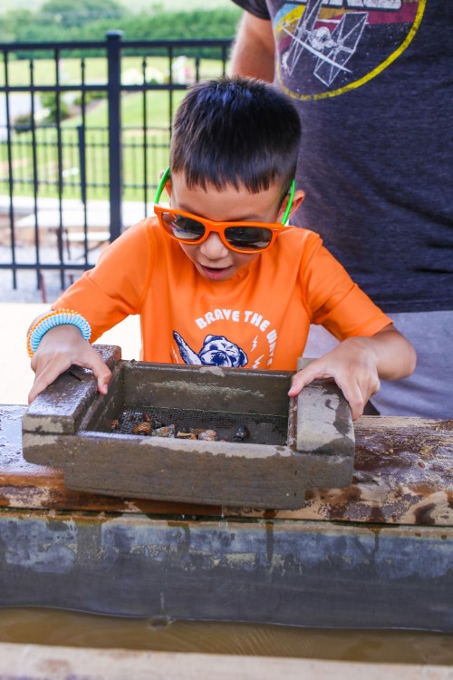 A young boy in sunglasses excitedly sifts through a container of water and sand, exploring for treasures.