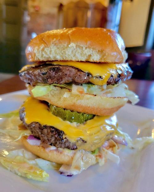 A towering cheeseburger with two beef patties, melted cheese, lettuce, pickles, and a soft bun on a plate.