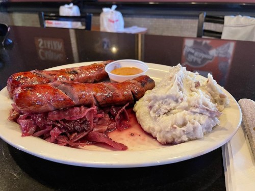 A plate with two grilled sausages, red cabbage, mashed potatoes, and a small cup of sauce on a black table.