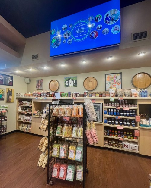 A store interior featuring shelves of products, a display rack of snacks, and a large screen showing promotional content.