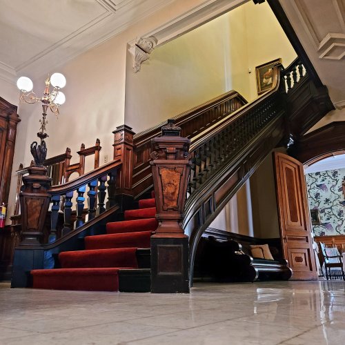 Elegant staircase with red carpet, wooden railings, and ornate chandeliers in a grand interior setting.