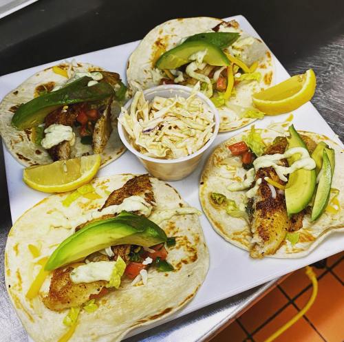 Four fish tacos with avocado, lettuce, and tomato, served with a side of coleslaw and lemon wedges.