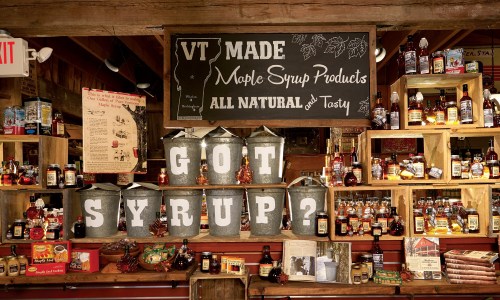 A rustic display of maple syrup products with metal buckets and shelves filled with syrup bottles, featuring a "Got Syrup?" sign.