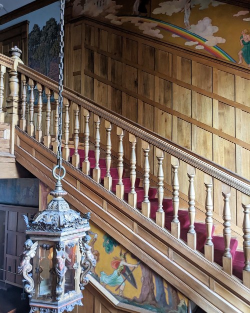 A wooden staircase with a red carpet, featuring ornate railings and vintage lantern, alongside colorful murals on the wall.