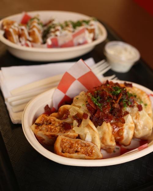 A bowl of dumplings topped with bacon and onions, served with a side of dipping sauce and chopsticks.