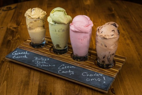 Four ice cream scoops in glasses on a wooden board, labeled: Caramel Crunch, Bailey's Irish Pistachio, Cotton Candy, Chocolate OMG!