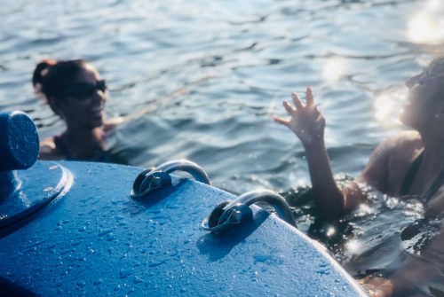 Two people chatting in the water near a blue boat, with sunlight reflecting on the surface.