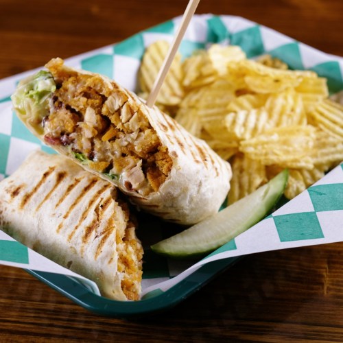 A grilled wrap filled with chicken and lettuce, served with potato chips and a pickle slice on a green-checkered tray.