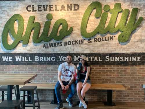 A man and woman sit on a bench in front of a green "Ohio City" mural, enjoying drinks in a casual setting.