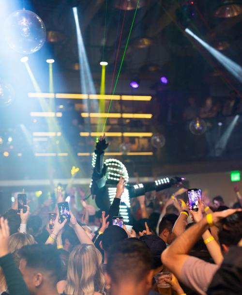 A lively nightclub scene with a performer in LED lights, surrounded by an energetic crowd holding phones.