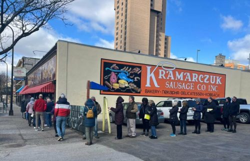 A long line of people waiting outside Kramarczuk Sausage Co. in a city setting, with a colorful sign on the building.