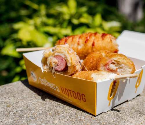 A box of crispy corn dogs with various toppings, resting on a stone surface with greenery in the background.