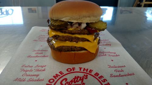 A towering cheeseburger with three patties, melted cheese, pickles, ketchup, and onions on a bun, served on branded paper.