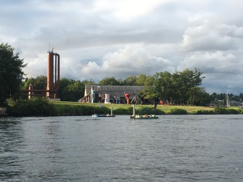 A view of a river with small boats, a grassy bank, and industrial buildings under a cloudy sky.