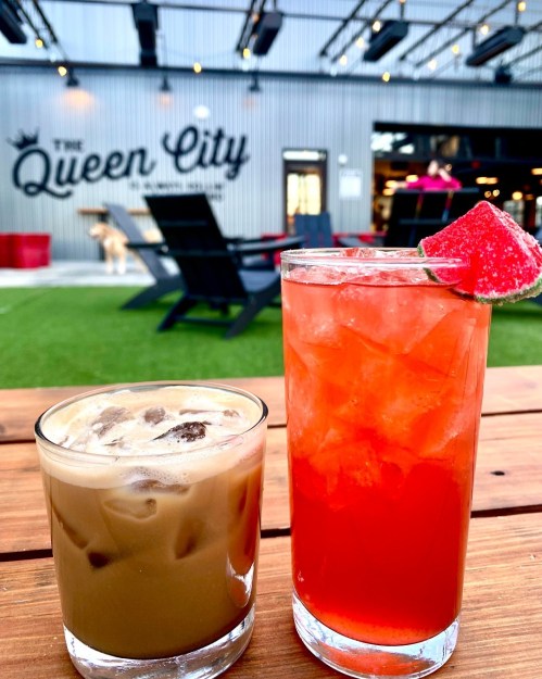 Two colorful drinks on a wooden table, with a lively outdoor setting featuring "The Queen City" in the background.