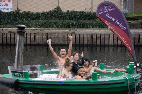 A group of five people enjoying a hot tub boat on a canal, smiling and posing with drinks. A promotional flag is visible.