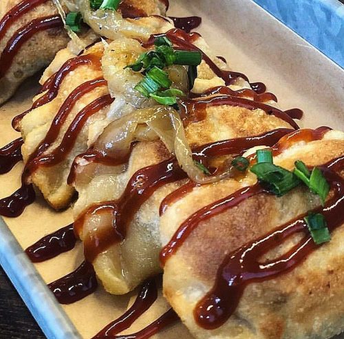A close-up of golden-brown dumplings drizzled with sauce and topped with green onions and sautéed onions.