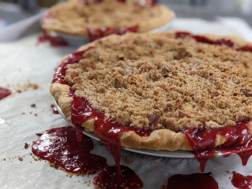 Two pies with crumb topping, overflowing with red fruit filling, sit on a sheet of parchment paper.