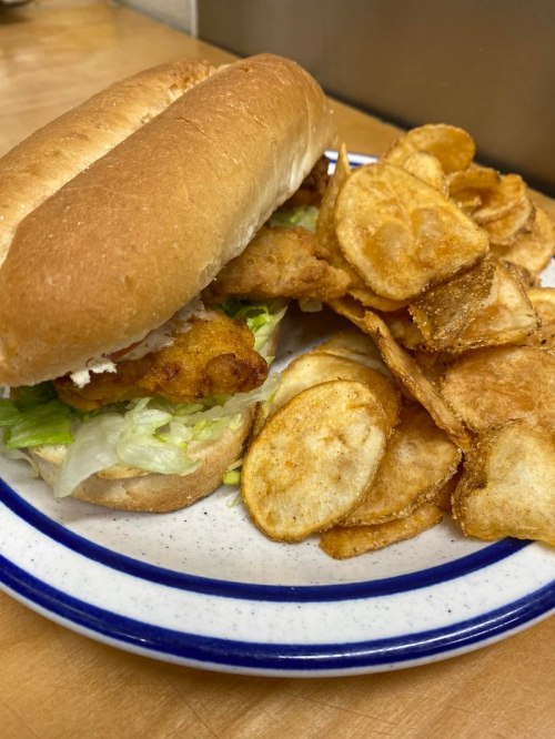 A sandwich with lettuce and fried fish on a bun, served with a side of crispy potato chips on a plate.