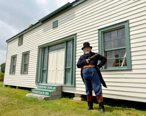 A man in period costume stands outside the Stagecoach Inn, holding a rifle, with a historic building in the background.