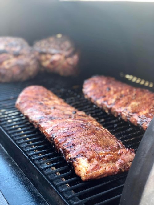 Two racks of ribs sizzling on a grill, with additional meat in the background.
