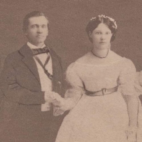 A vintage black-and-white photo of a man and woman holding hands, both dressed in formal attire from the 19th century.