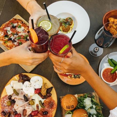 A group cheers with colorful cocktails over a table filled with pizza, appetizers, and a charcuterie board.