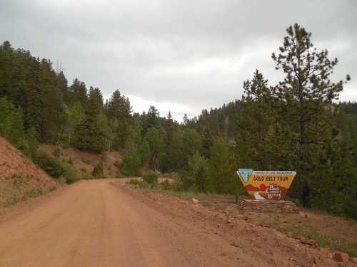 A dirt road winds through a forested area, with a sign for the Gold Belt Tour visible on the right.