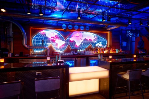 A vibrant bar with a glowing world map backdrop, colorful lighting, and a sleek counter with bar stools.
