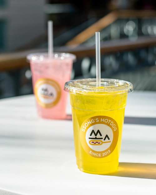 Two colorful drinks in clear cups with straws, one yellow and one pink, on a table with a logo for Ssong's Hotdog.