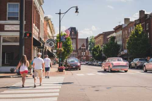 A sunny street scene with people walking, vintage cars, and historic buildings lining the road.