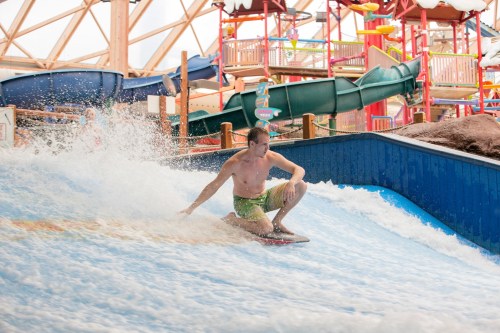 A man surfing on a wave at an indoor water park, surrounded by colorful slides and attractions.